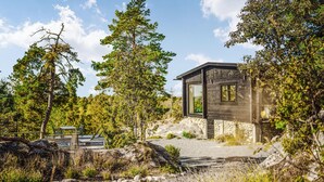 Une cabane moderne, dotée de fondations en pierre, entourée d’une zone boisée et accessible par un chemin de gravier.