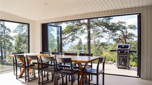 Un coin repas avec une table et des chaises en bois, de grandes fenêtres donnant sur la verdure et un barbecue à l’extérieur.