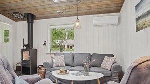 A cozy living room with a grey sofa, two armchairs, a round coffee table, a wood-burning stove, a hanging light fixture, and a window with a view of greenery. - Amazing home in Brovst with kitchen (Brovst)