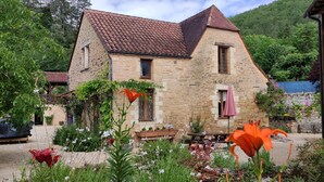 Una casa de piedra con techo de tejas, rodeada de un jardín con diversas plantas y flores.