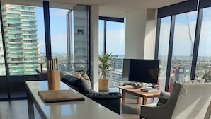 A modern living room with a large window, a flat-screen TV, and a dining area. - Top Floor Stunning Views (South Melbourne)