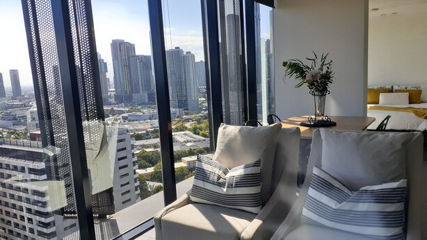 A modern living room with a city view, featuring a sofa and armchair with striped pillows, a dining table, and a potted plant. - Top Floor Stunning Views (South Melbourne)