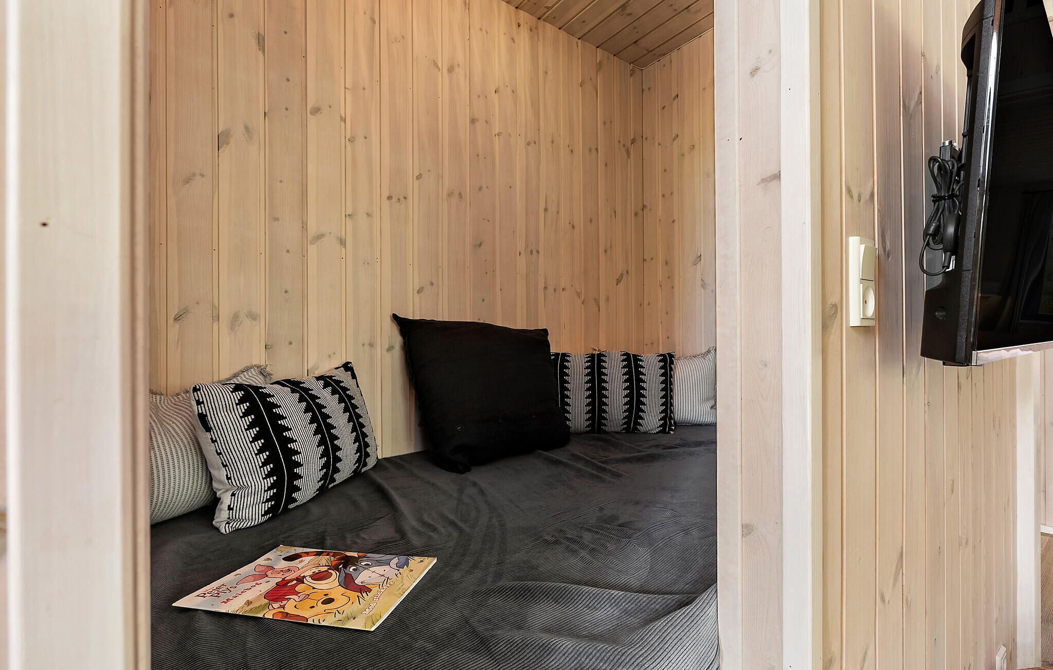 Quarto pequeno e acolhedor com teto e paredes de ripas de madeira, uma cama de casal com colchão cinza escuro e almofadas estampadas, e uma TV de tela plana fixada na parede.
