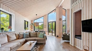 A spacious living area with a large sofa, a coffee table, and a television. There are large windows and a view of the outdoors. - Amazing home in Sydals with sauna (Sydals)