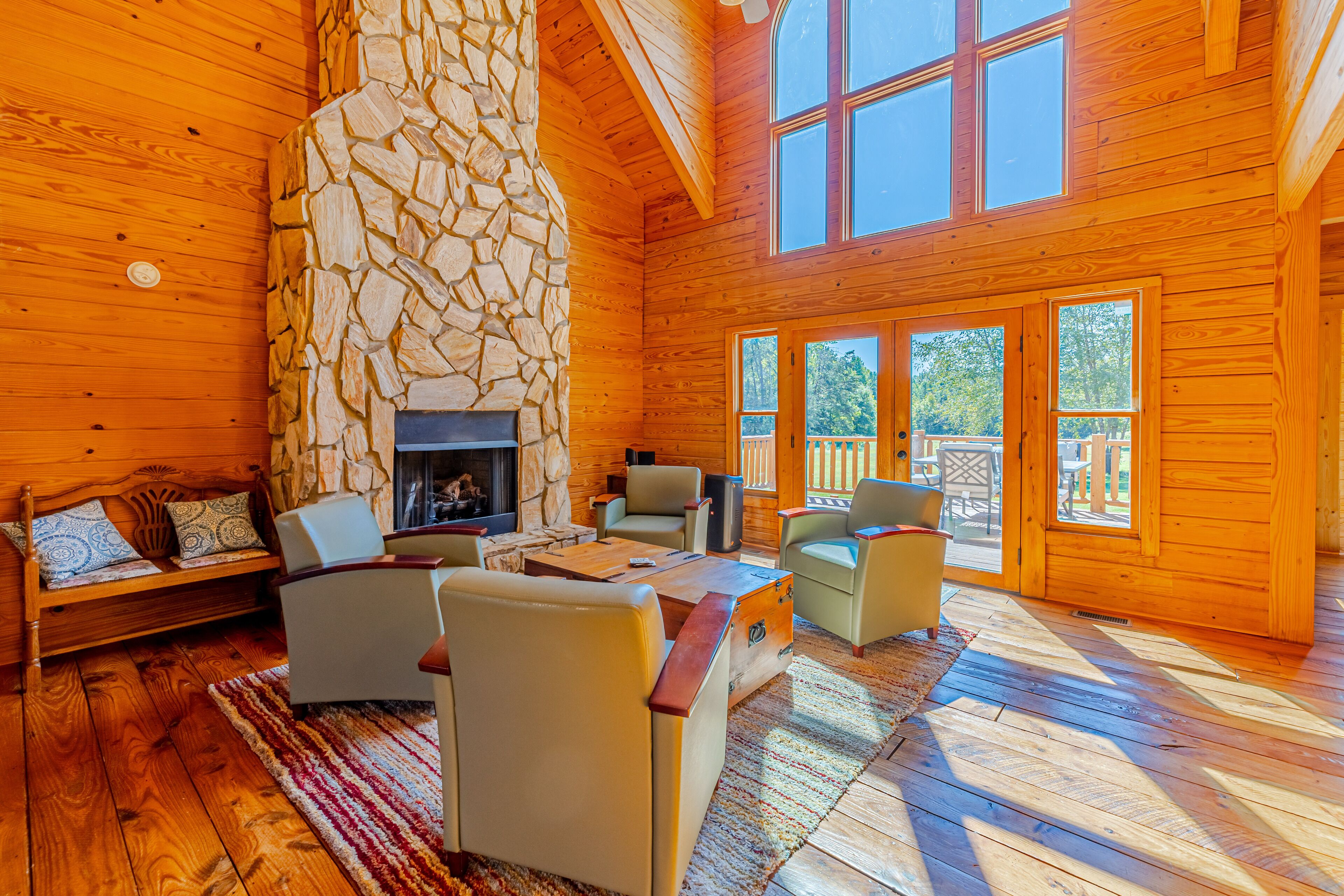 A log cabin interior with a stone fireplace, wooden furniture, and large windows.