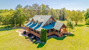 A large, two-story log cabin with a wrap-around porch and multiple decks, surrounded by a well-maintained lawn and dense forest.