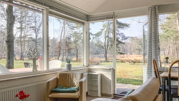 Ein Wintergarten mit großen Fenstern, ein gepolsterter Sessel, ein kleiner Tisch mit Blumenvase und Blick auf einen bewaldeten Bereich.