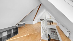 Une pièce mansardée spacieuse avec un plafond en pente, un plancher en bois, une rambarde blanche et un petit coin salon agrémenté d’un coussin à motifs.