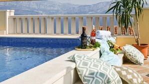A poolside area with a view of mountains, a white railing, and a table with bottles and fruits. - Amazing home in Postira (Postira)