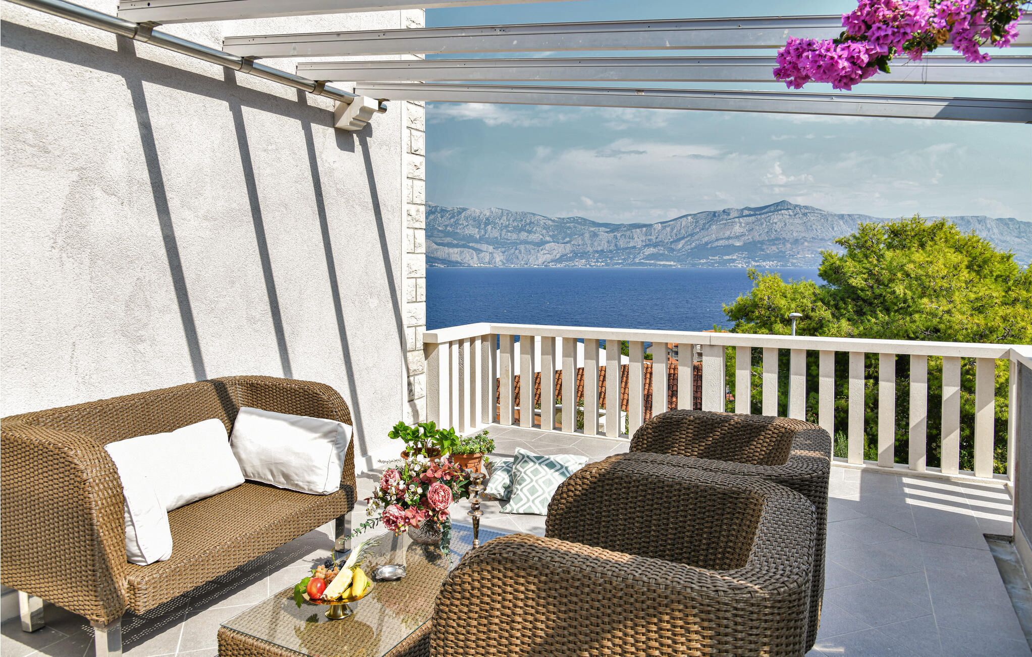 A balcony with wicker furniture, a glass table with fruit, and a view of the sea and mountains.