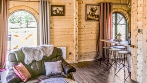 Interior acogedor de una cabaña de madera con un sofá de cuero, una mesa de comedor con taburetes y dos ventanas arqueadas con cortinas.
