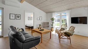 Een moderne woonkamer met een zwarte leren bank, een houten salontafel, rieten fauteuils en een flatscreen televisie aan de muur.