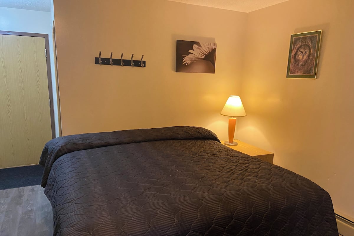 A hotel room with a bed, a nightstand with a lamp, a wall-mounted coat rack, and two framed pictures.