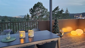 Una terraza de madera con mesa y sillas, iluminación exterior y vista a un paisaje urbano al fondo.