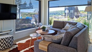 Una sala moderna con un sofá grande, una mesa de centro y un televisor de pantalla plana. Amplias ventanas ofrecen vistas al exterior.