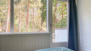 Une chambre avec une fenêtre donnant sur une forêt, un mur en briques blanches et un lit recouvert d’une couverture verte.