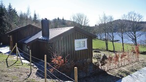 A small wooden cabin with a chimney, a fenced yard, and a lake in the background. - Awesome home in Farsund with house sea view (Farsund)