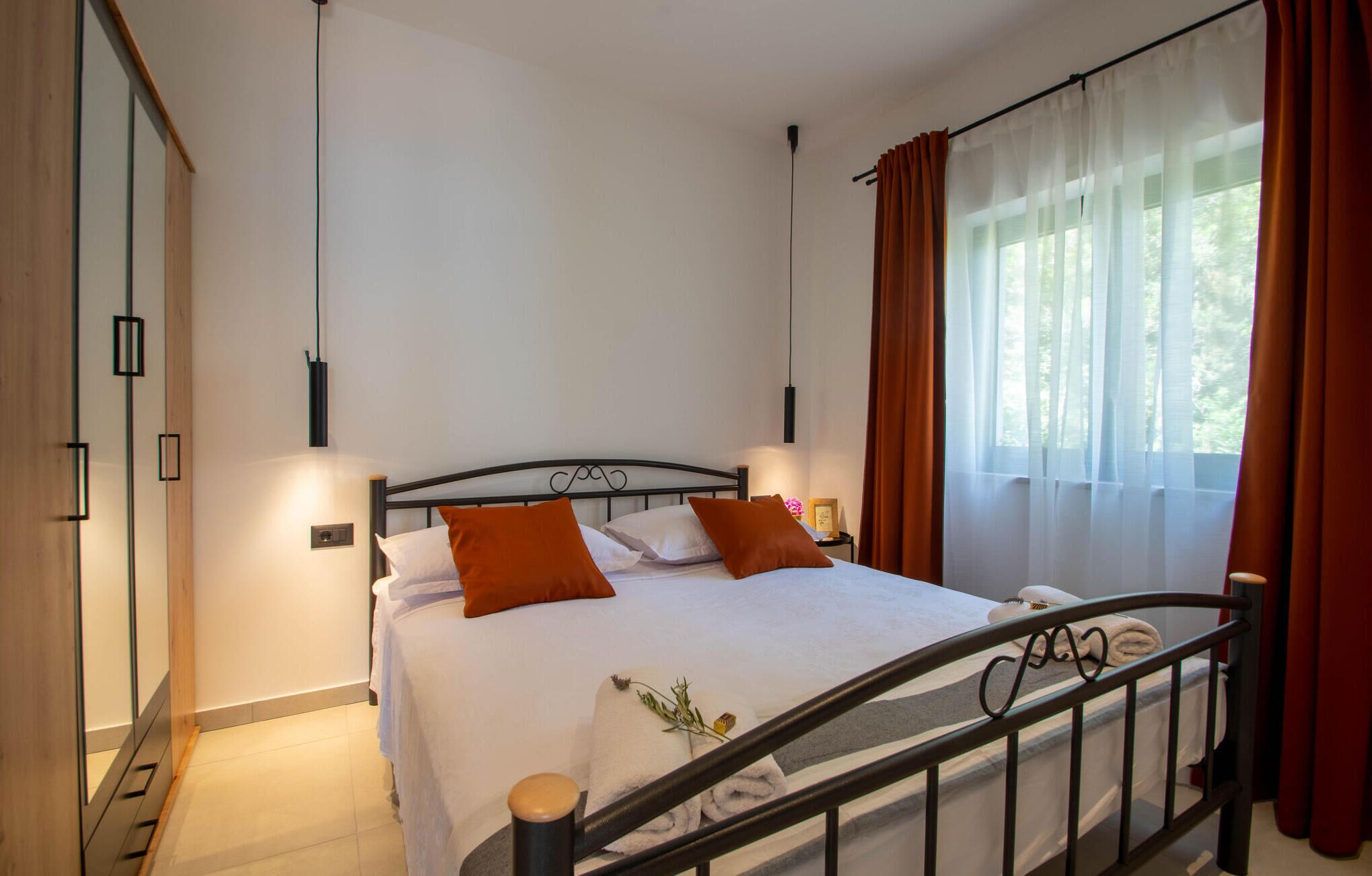 A neatly made bed with white linens and orange pillows, a bedside table with a lamp, and a window with sheer curtains and a view of greenery.
