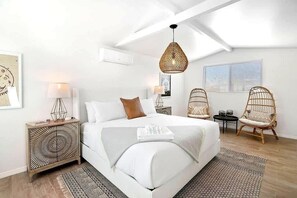 A modern bedroom with a large bed, two bedside tables, a hanging light fixture, wicker chairs, and a window with blinds.