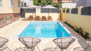 Una piscina rectangular con agua azul cristalina, rodeada de baldosas de piedra. Hay cuatro sillas de mimbre para tomar el sol y dos sombrillas que dan sombra.