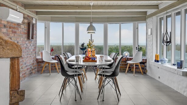Een eetruimte met een tafel gedekt voor zes personen, omringd door stoelen, een hangende lamp en uitzicht op het landschap door grote ramen.