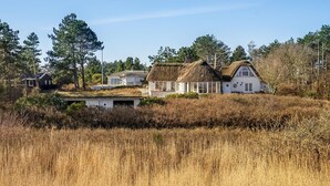 Een rieten huis met een witte gevel, omringd door hoog gras en bomen.