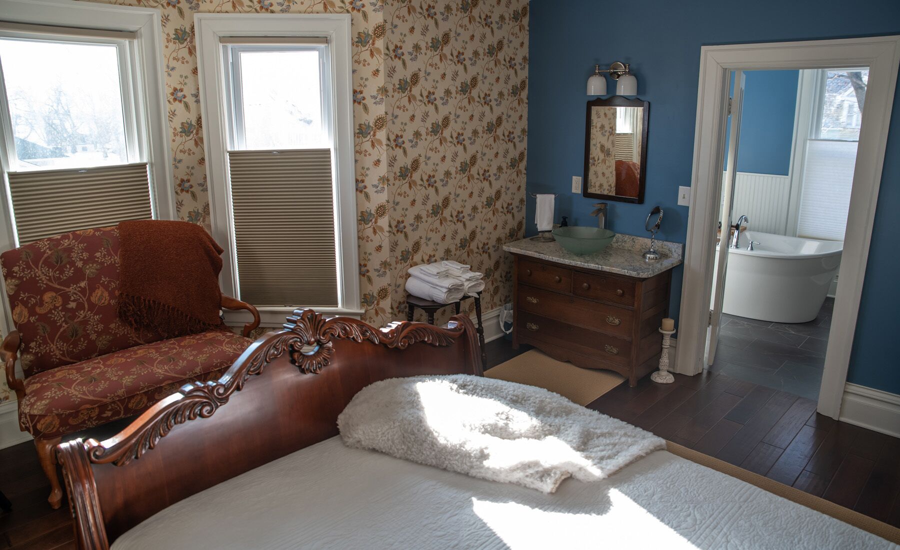 A bedroom with a large bed, a wooden chair, a dresser, a mirror, and a bathroom with a sink and bathtub.