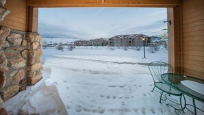 Un patio cubierto de nieve con un muro de piedra, una mesa y una silla de metal verde, y vistas a una zona residencial con edificios al fondo.