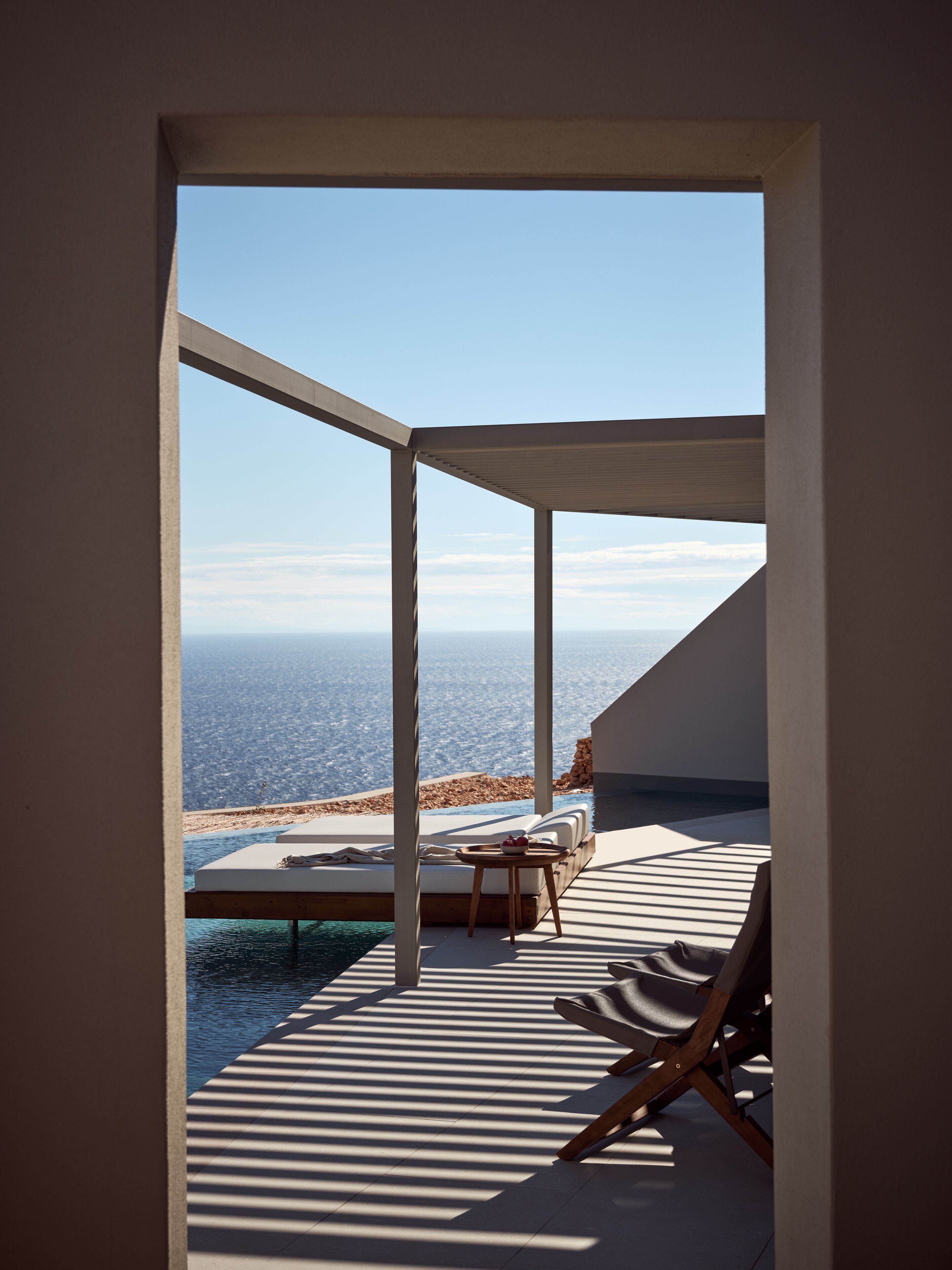 A modern outdoor lounge area with a view of the sea, featuring a pool, lounge chairs, and a small table.