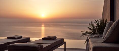 A sunlit terrace with a view of the ocean at sunset, featuring lounge chairs and a potted plant.