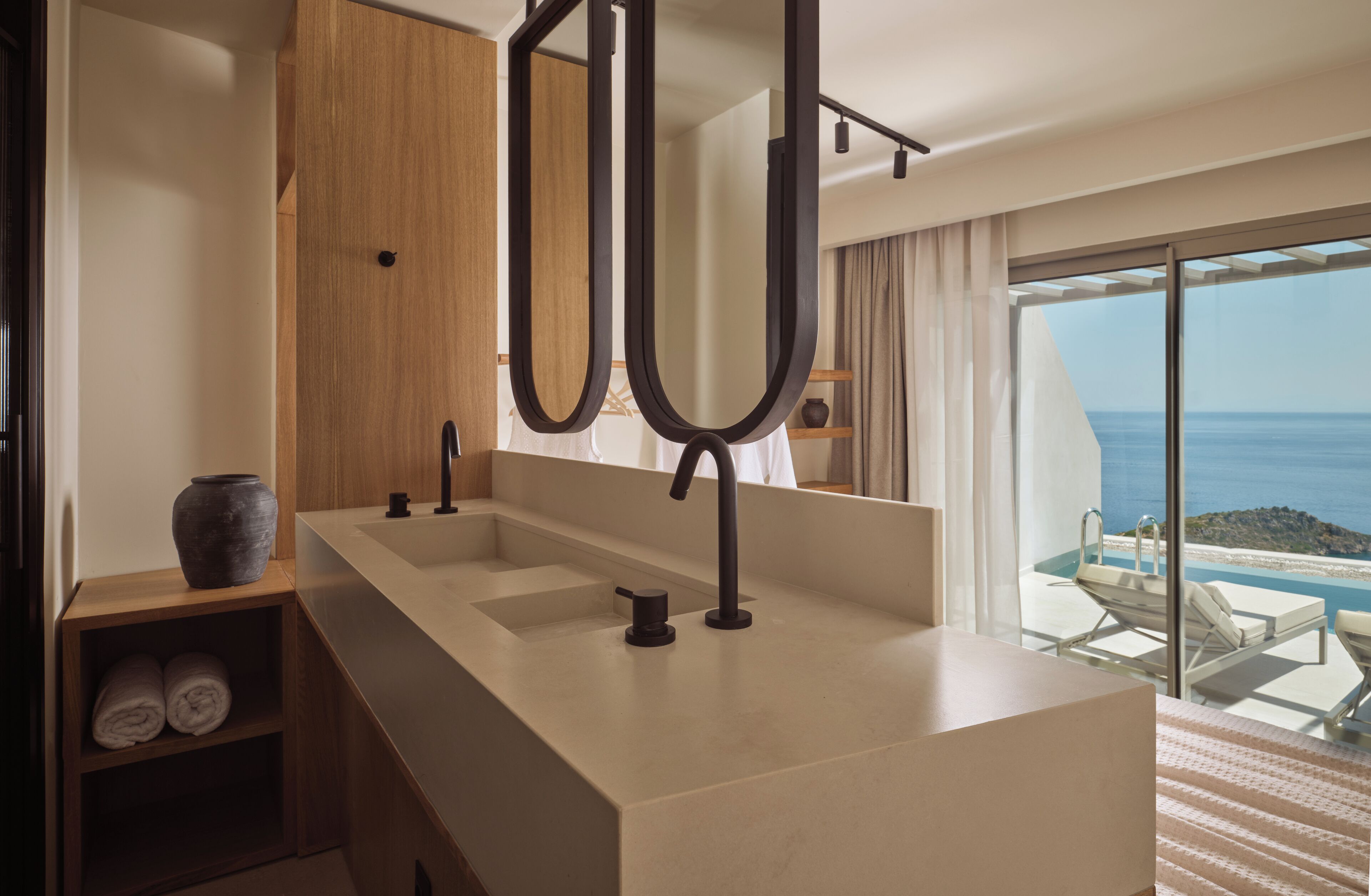Une salle de bain moderne avec un grand miroir, un meuble en bois et une vue sur l’océan grâce à une porte-fenêtre coulissante.