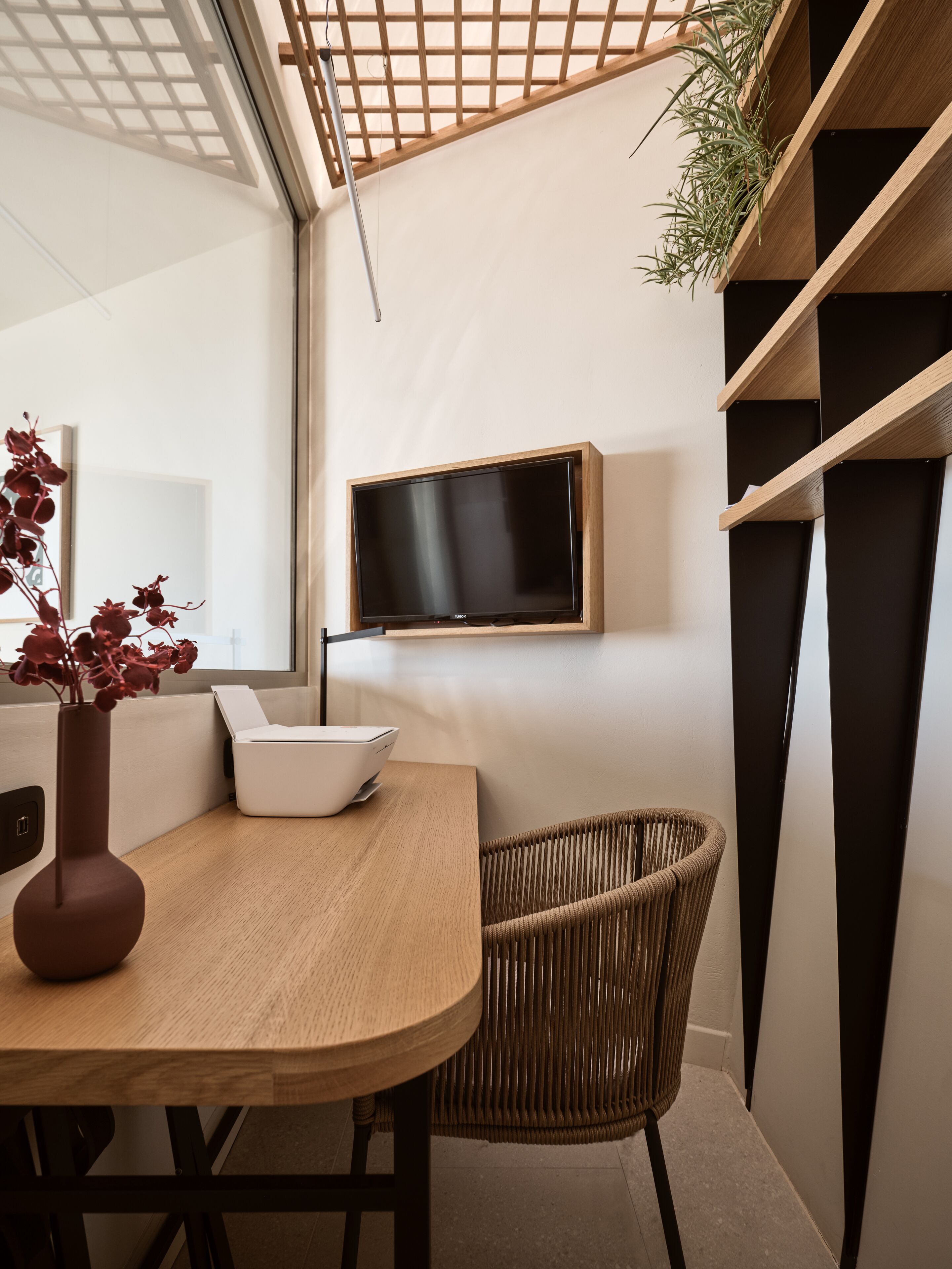 Un intérieur moderne avec une table en bois, une chaise en osier, un vase blanc orné de fleurs rouges, un téléviseur à écran plat fixé au mur et une étagère noire.