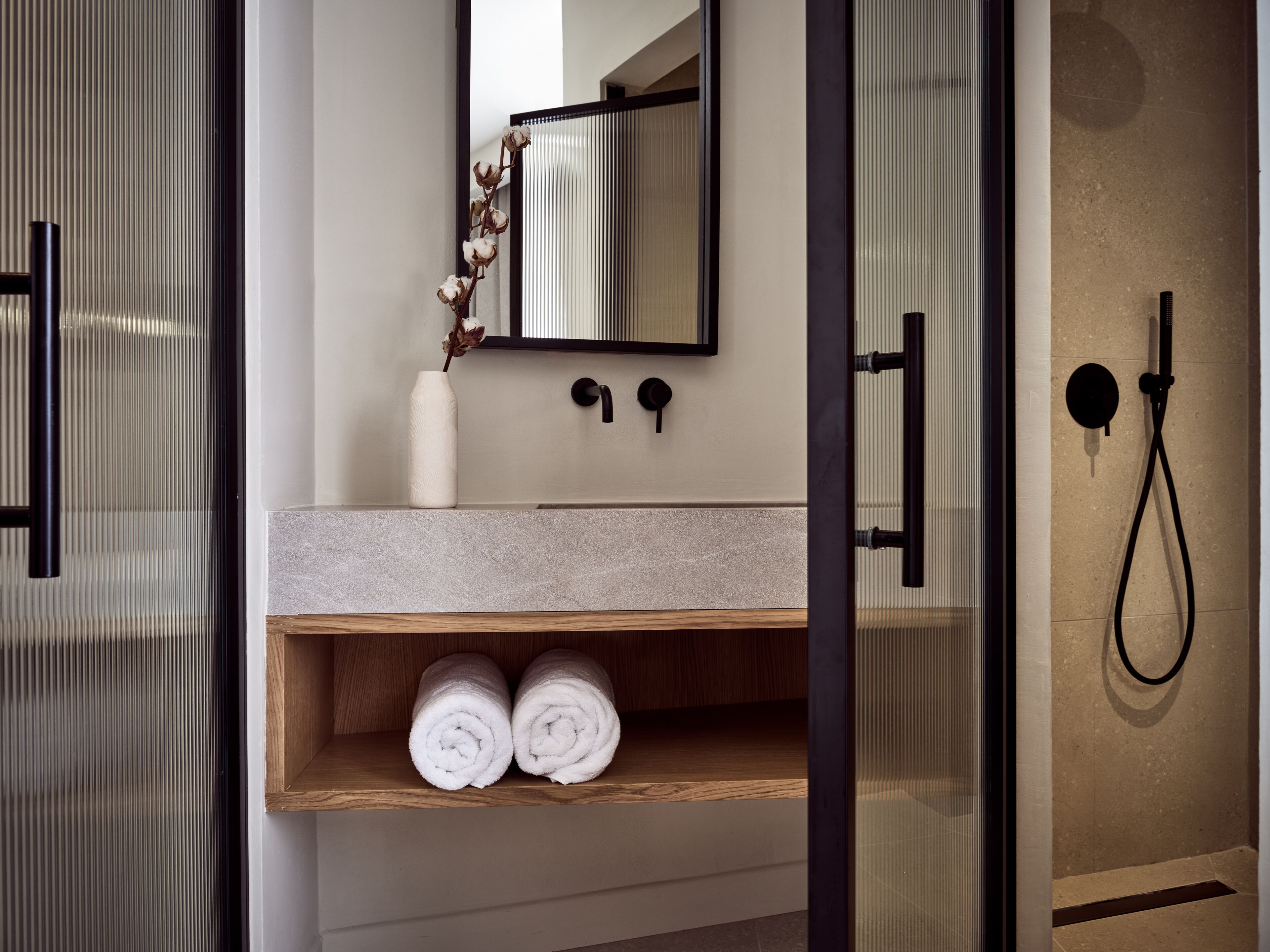 Een moderne badkamer met een marmeren aanrecht, een houten plank met opgerolde handdoeken, een witte vaas met gedroogde bloemen, een spiegel en een zwarte douchekop.