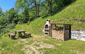 Un’ambientazione esterna rustica con una griglia per barbecue in pietra, un tavolo e una panchina in legno, e una pila di legna.