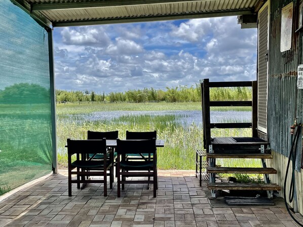 A covered patio with a table and chairs, overlooking a water body and greenery.