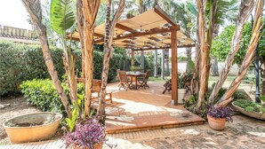 A covered outdoor patio with a wooden table and chairs, surrounded by tropical plants and a pergola structure. - 1 bedroom lovely home in Siracusa (Siracusa)
