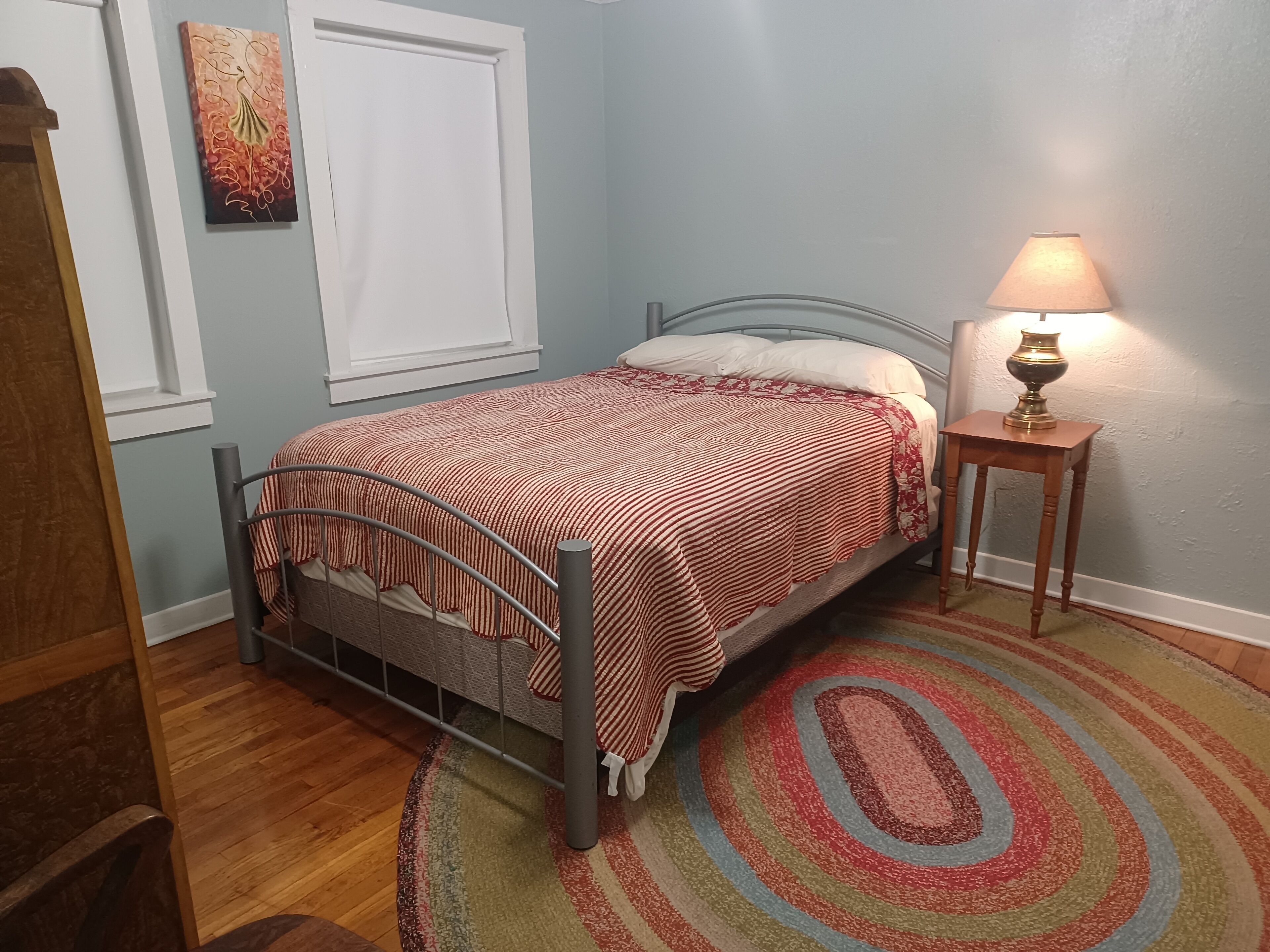 Een slaapkamer met een bed, een nachtkastje met een lamp, een raam met een witte jaloezie en een kleurrijk vloerkleed.