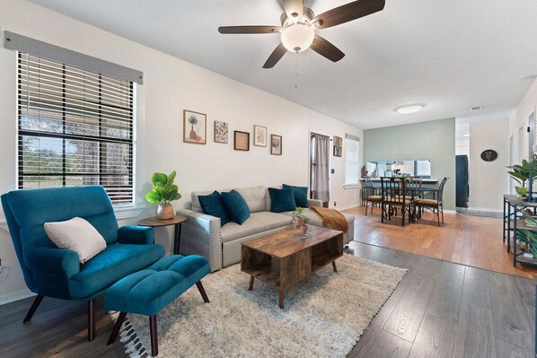 A living room with a teal armchair, a wooden coffee table, and a sofa with blue cushions. There is a ceiling fan, a dining area with a table and chairs, and a kitchen in the background.