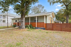 A single-story house with a porch, a fenced yard, and a large tree in front.