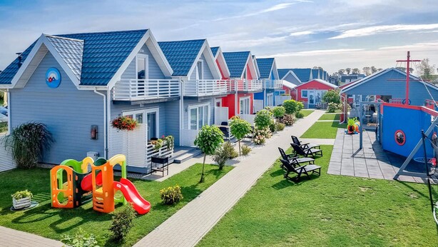 Une rangée de maisons mobiles aux couleurs vives, avec une aire de jeux, une structure en forme de bateau et une allée pavée.