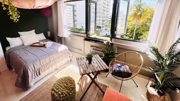 A modern bedroom with a large bed, a small table, a chair, and a view of a cityscape through large windows.