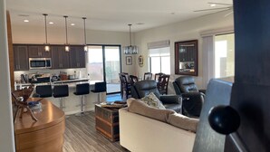 A modern living room with a sofa, a dining area with chairs, a kitchen with dark cabinets and a microwave, and a large window. - Colorado River Getaway, Resort Pool, Waterslide & Mountain Views (Fort Mohave)