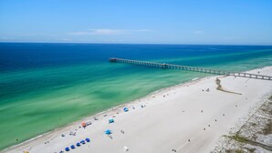 An aerial view of a beach with a pier extending into the ocean, sandy beach, and clear blue water.
