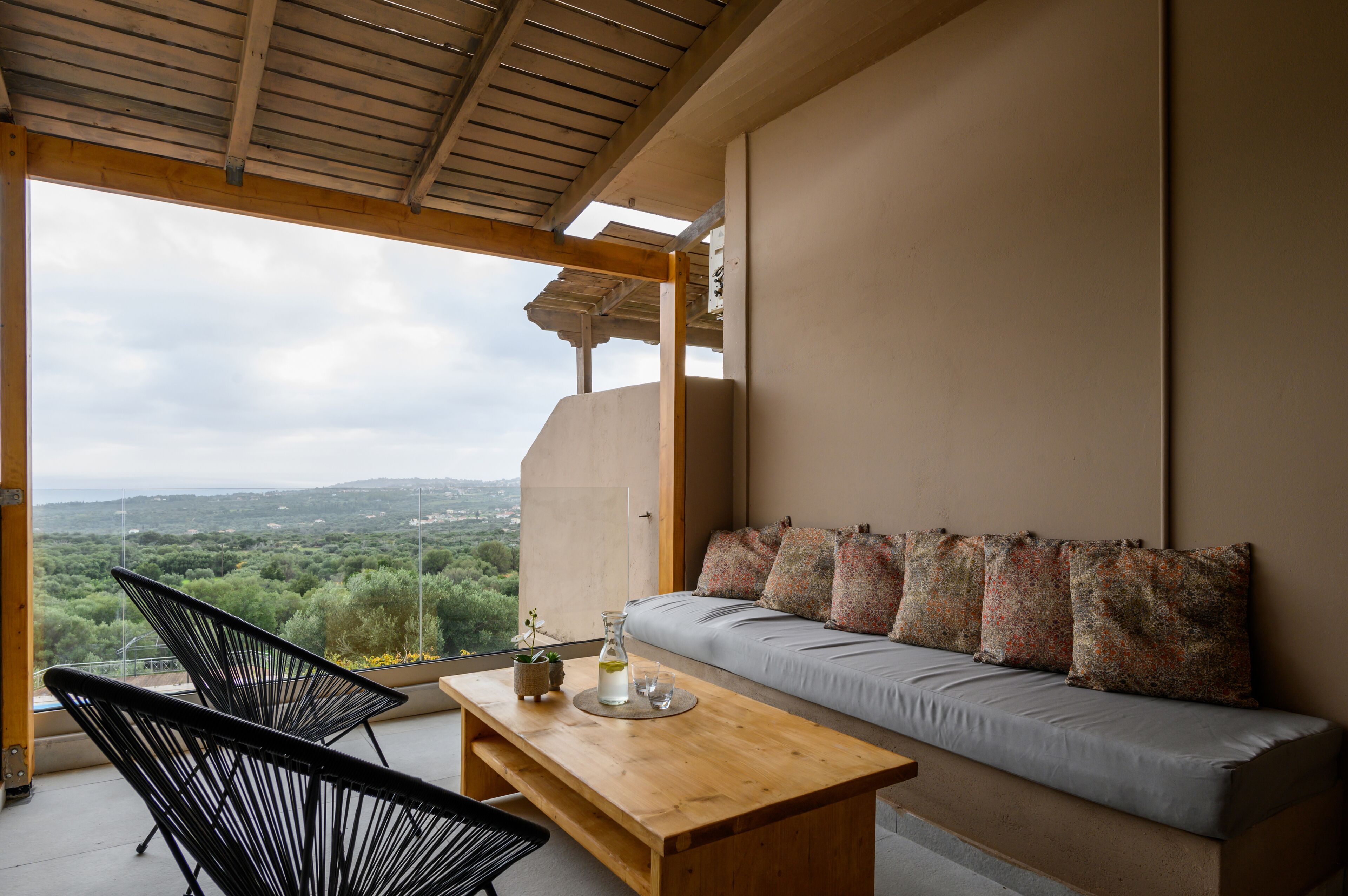 A balcony with a sofa and chairs, a wooden table, and a view of a green landscape.