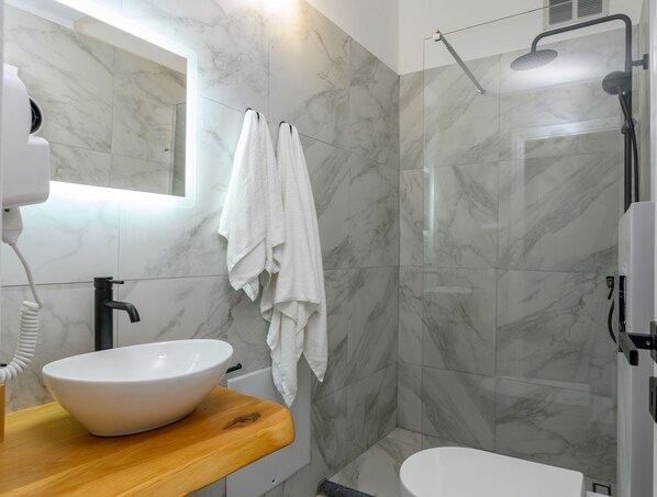 Un baño moderno con salpicadero de mármol, encimera de madera, lavabo blanco, toallero y zona de ducha con mampara de vidrio.
