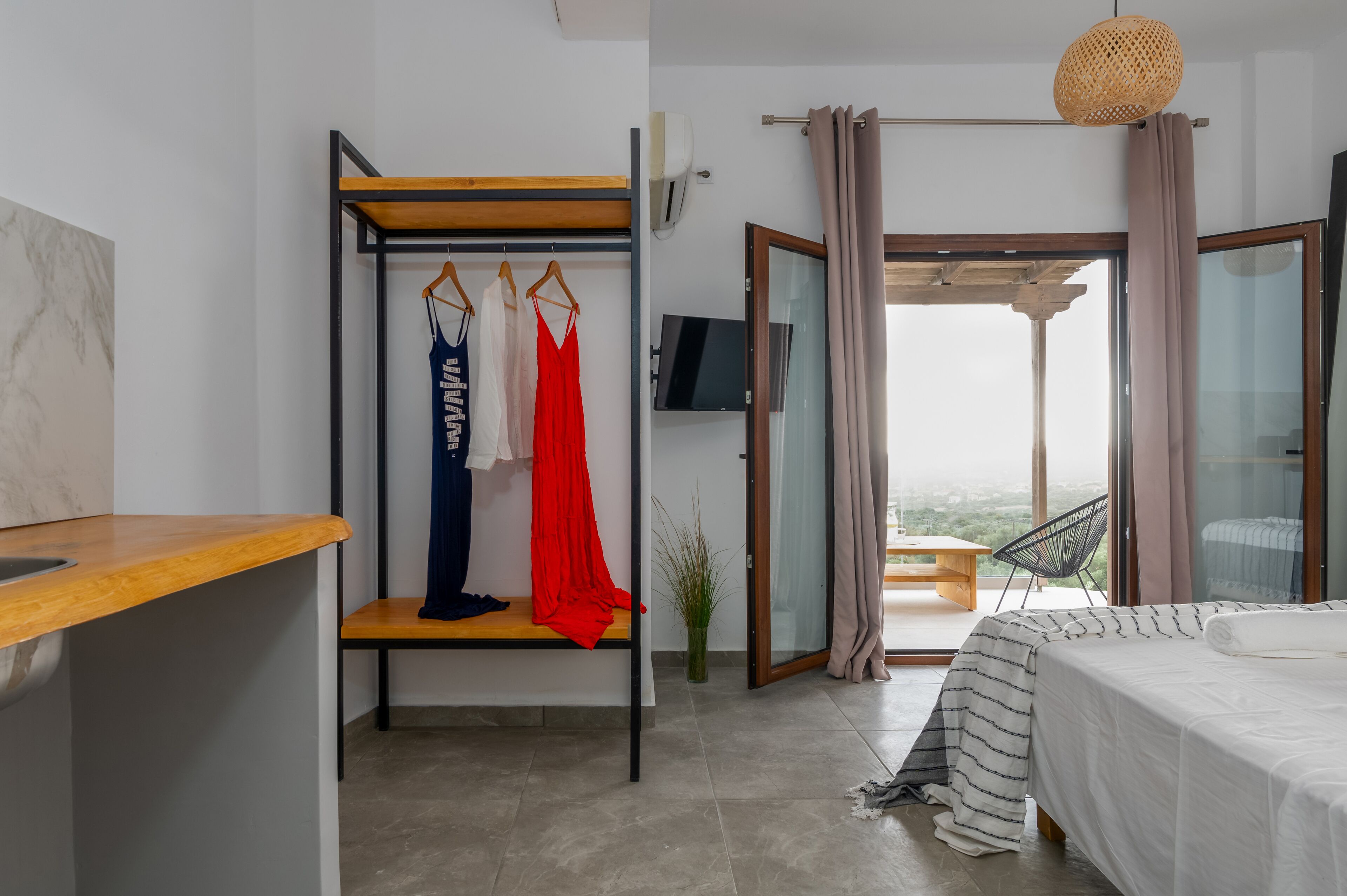 Une chambre à coucher avec un lit, un banc en bois, un portant à vêtements avec des vêtements suspendus, une télévision et une vue sur l’extérieur, visible à travers une porte ouverte.