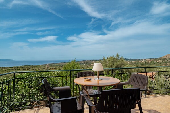 Una terraza con una mesa redonda y cuatro sillas, una lámpara de mesa, y vistas al mar y las colinas.