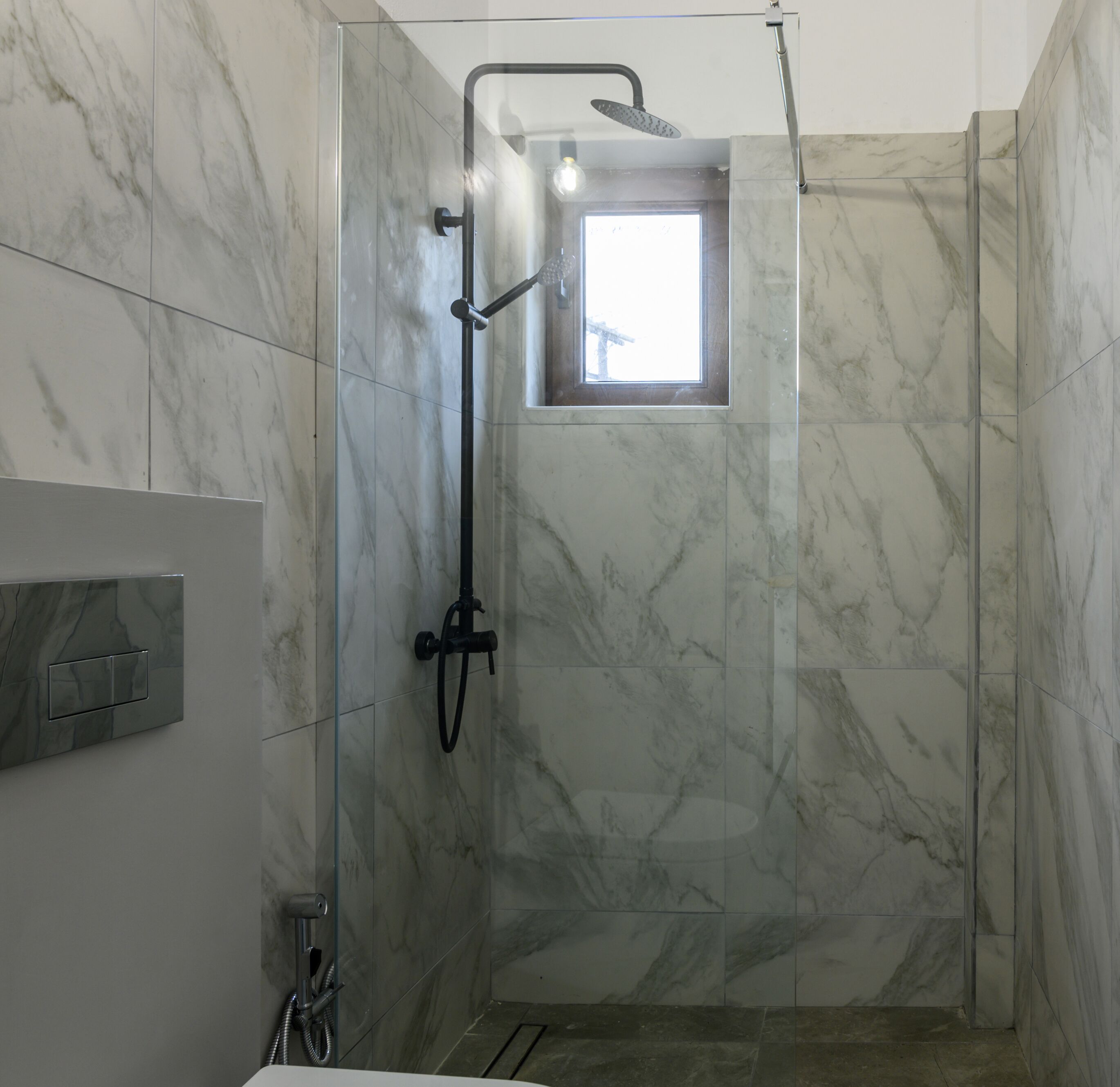 A glass-enclosed shower with a handheld showerhead and a fixed showerhead, marble walls, and a small window.