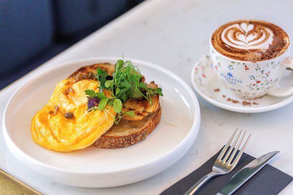Un plato de desayuno con tostadas, huevos revueltos y vegetales verdes, junto a una taza de café con arte de latte sobre una mesa.