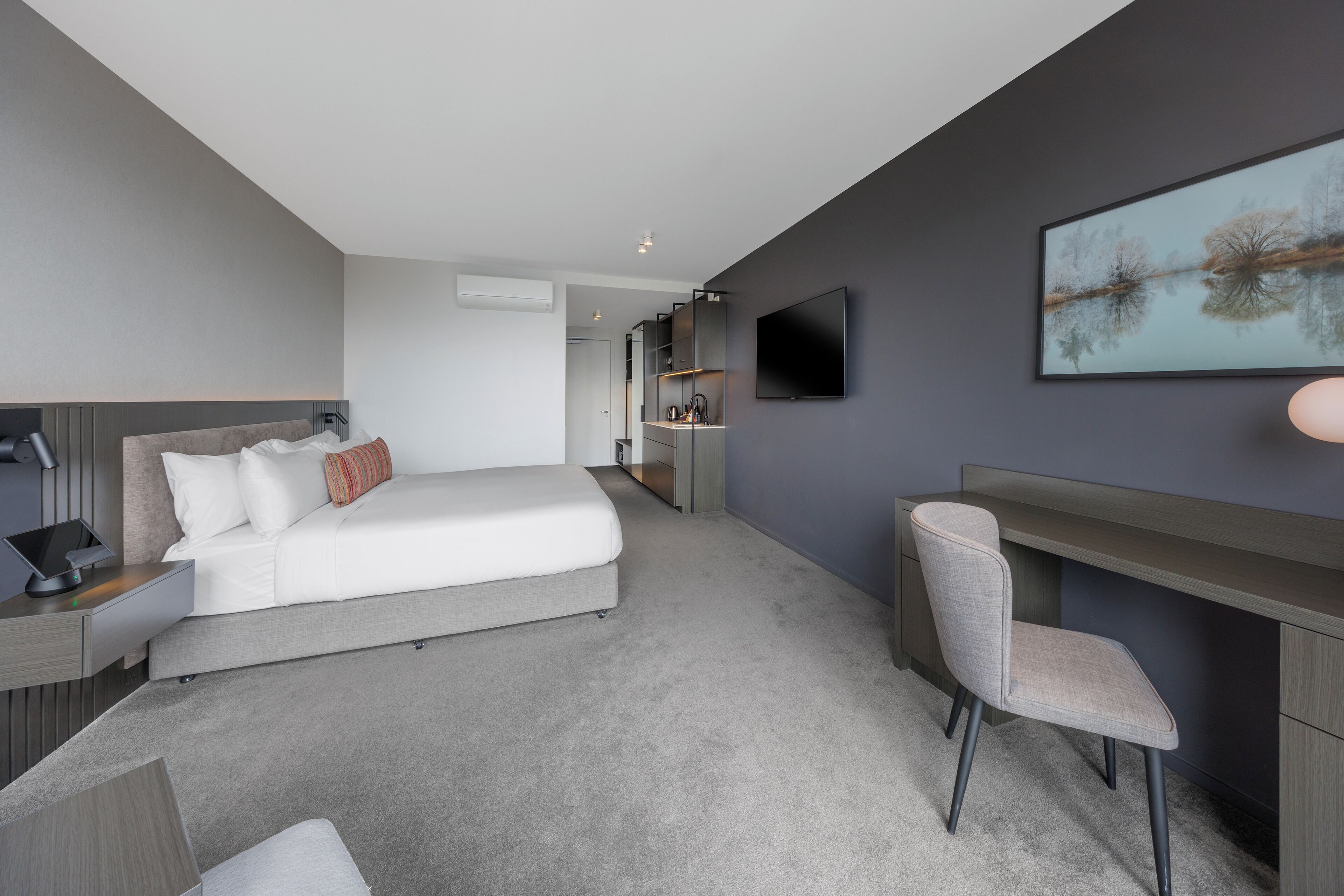 Ein modernes Hotelzimmer mit Bett, Nachttisch, Schreibtisch, Stuhl, Fernseher und einem Bild an der Wand.